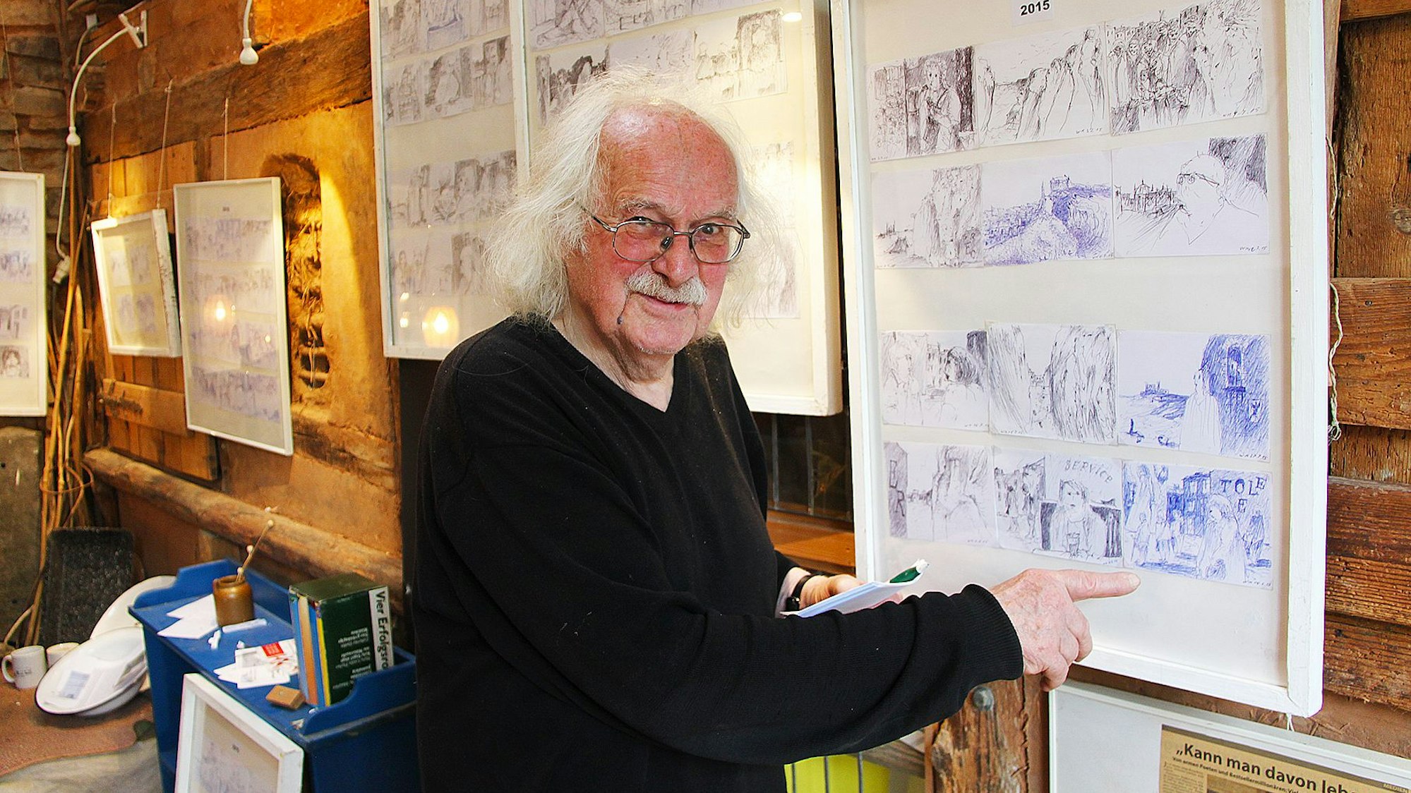 Wolfgang Martens steht vor kleinen Skizzen, die in Gruppen gerahmt sind.