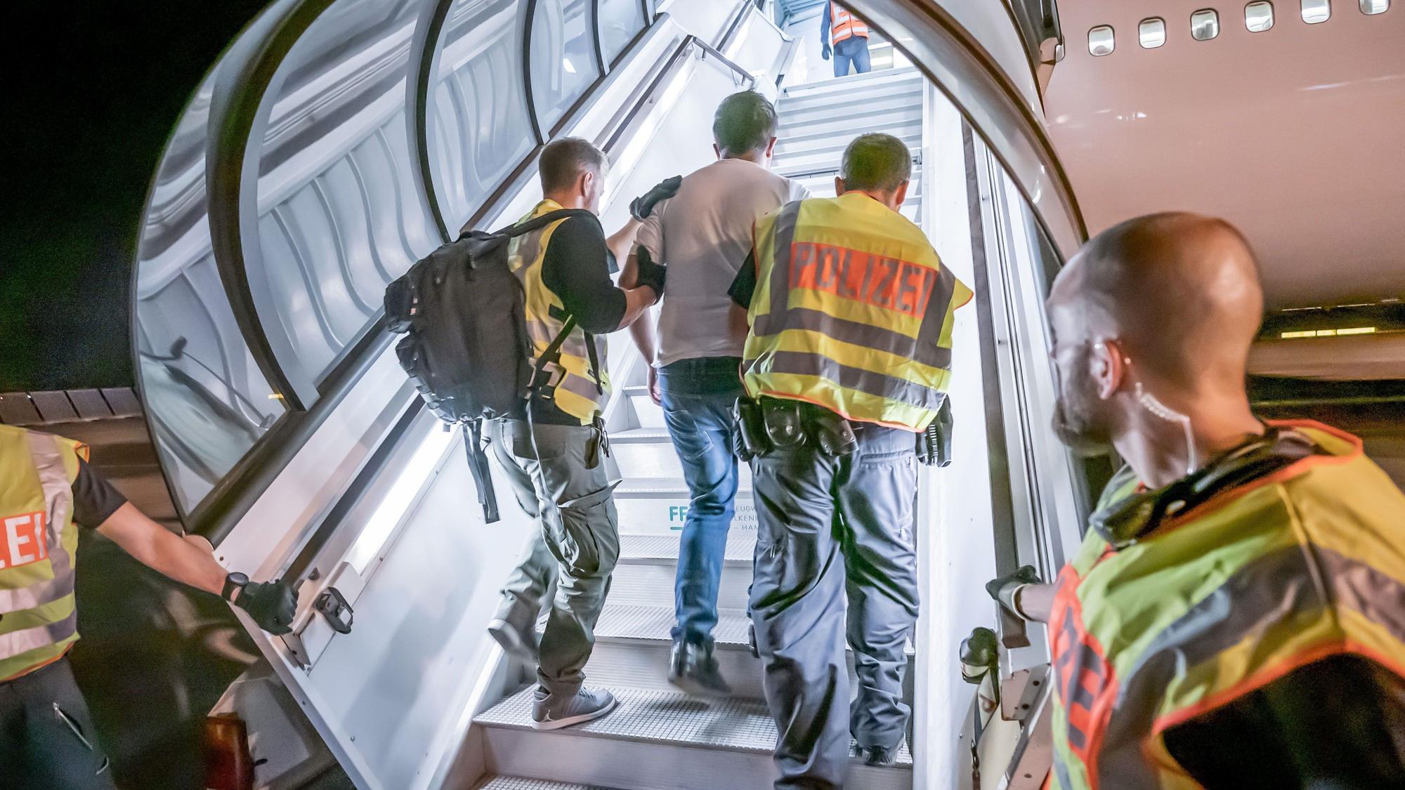 Das Bild zeigt einen Mann, der von Polizisten in ein Flugzeug begleitet wird, damit er mithilfe des Flugs abgeschoben werden kann.