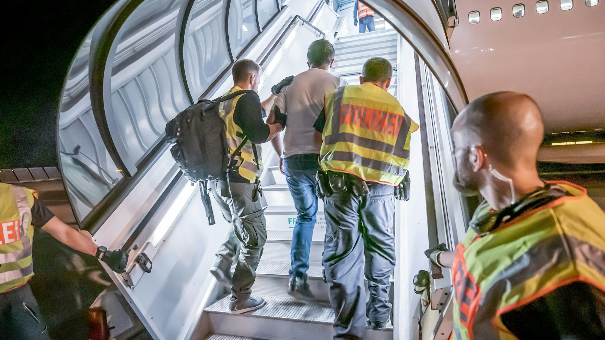 Das Bild zeigt einen Mann, der von Polizisten in ein Flugzeug begleitet wird, damit er mithilfe des Flugs abgeschoben werden kann.