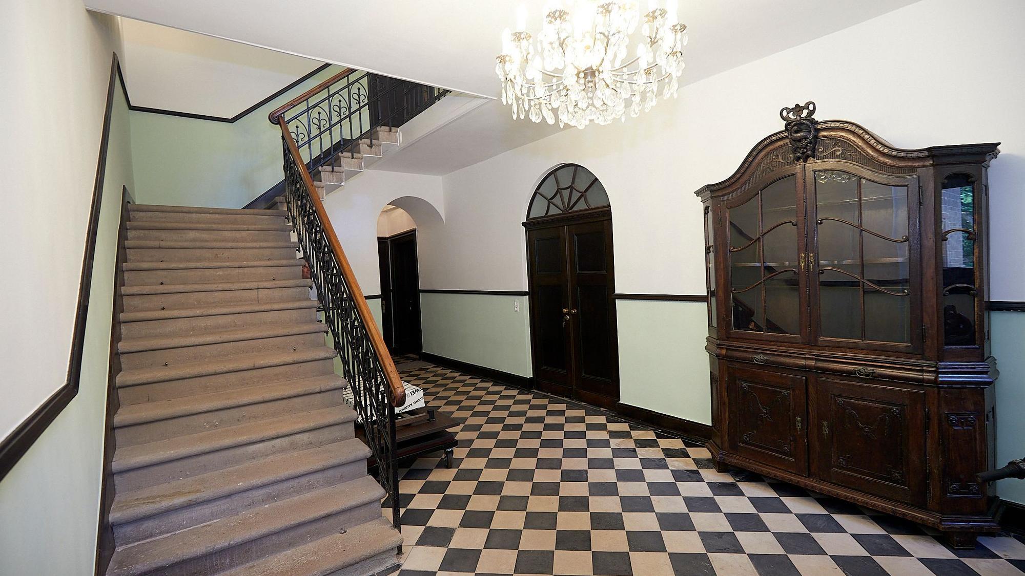 Vom Flur aus führt eine Treppe ins obere Geschoss. An einer Wand steht ein aufwendig gestalteter Schrank, an der Decke hängt ein Kronleuchter.