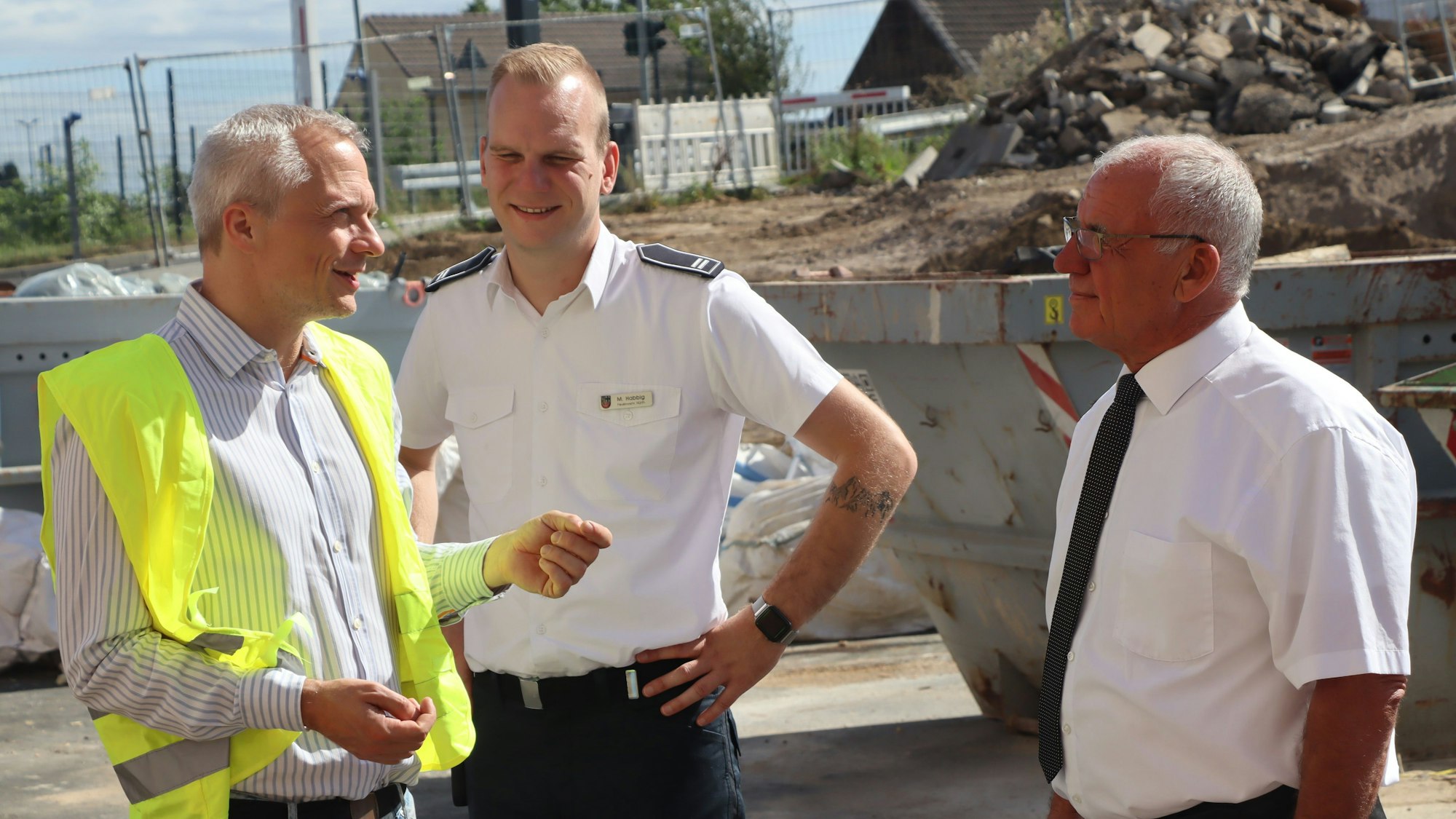 Ein Mann in Warnweste und zwei Männer in Feuerwehr-Uniform auf der Baustelle.