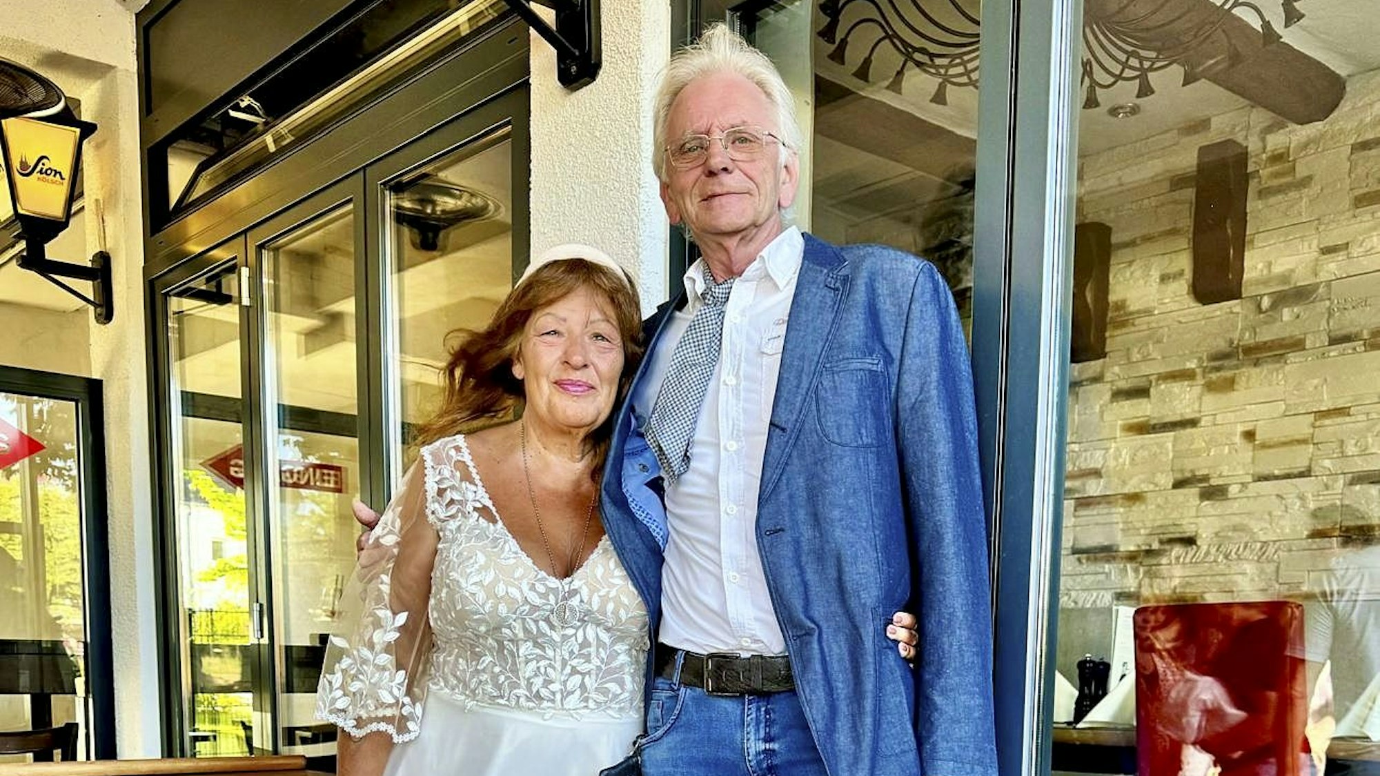 Eine ältere Frau im Brautkleid legt ihren Arm um einen größeren, älteren Herrn im Anzug.