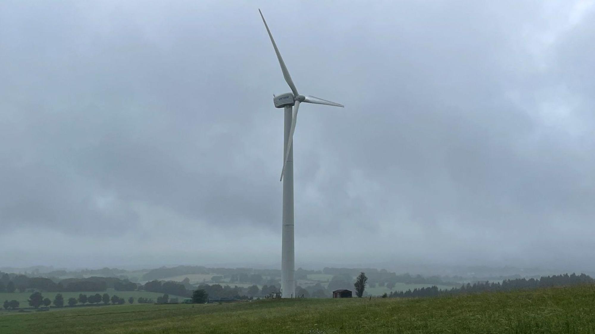 Ein einzelnes Windrad steht auf einer grünen Wiese vor wolkenverhangenem Himmel, im Hintergrund sind grüne Hügel und Wälder zu sehen.