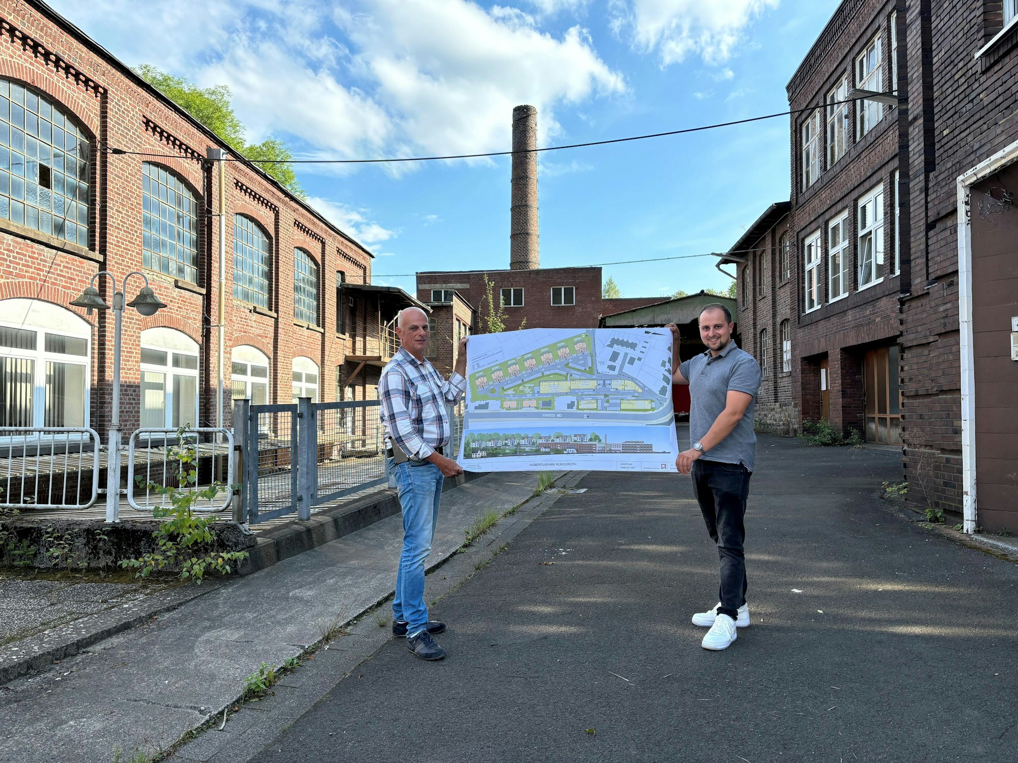 Zwei Männer stehen auf dem Hof einer alten Fabrik und halten gemeinsam die Pläne für die Neuentwicklung des Areals in Händen.