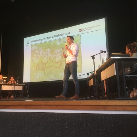 Ein Mann in Jeans und weißem Hemd steht auf Bühne des Theaters am Park, er spricht in ein Mikrofon. An der Wand hinter ihm ist eine Karte mit Windkraft-Potenzialflächen in Eitorf zu sehen.