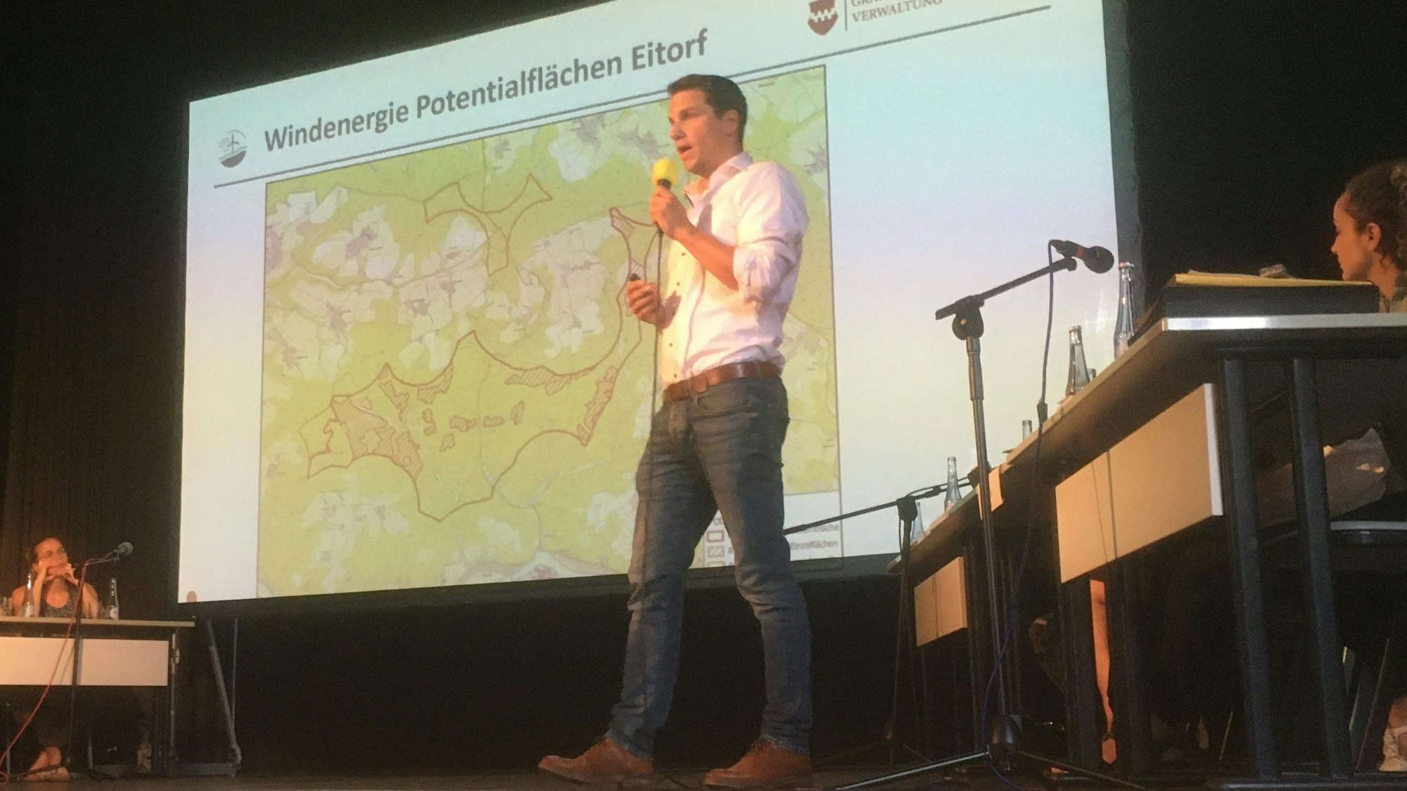 Ein Mann in Jeans und weißem Hemd steht auf Bühne des Theaters am Park, er spricht in ein Mikrofon. An der Wand hinter ihm ist eine Karte mit Windkraft-Potenzialflächen in Eitorf zu sehen.