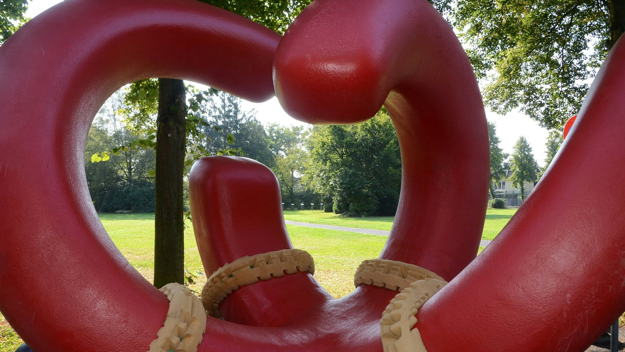 Eine moderne rote Skulptur steht vor einer parkähnlichen Wiese.