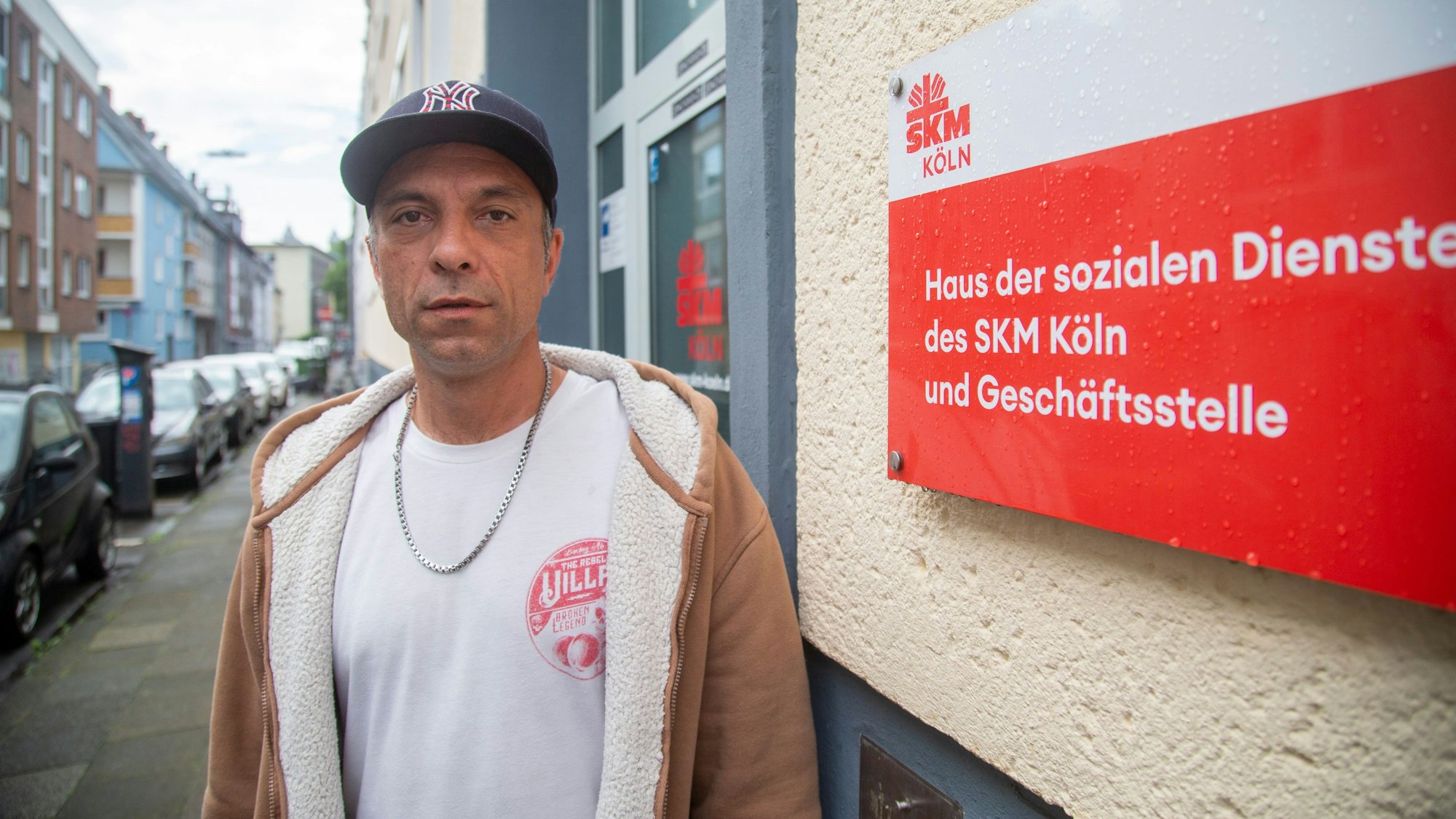 An Mann steht neben einem Haus, an dem ein Schild angebracht ist, auf dem steht „Haus der sozialen Dienste des SKM Köln und Geschäftsstelle“.