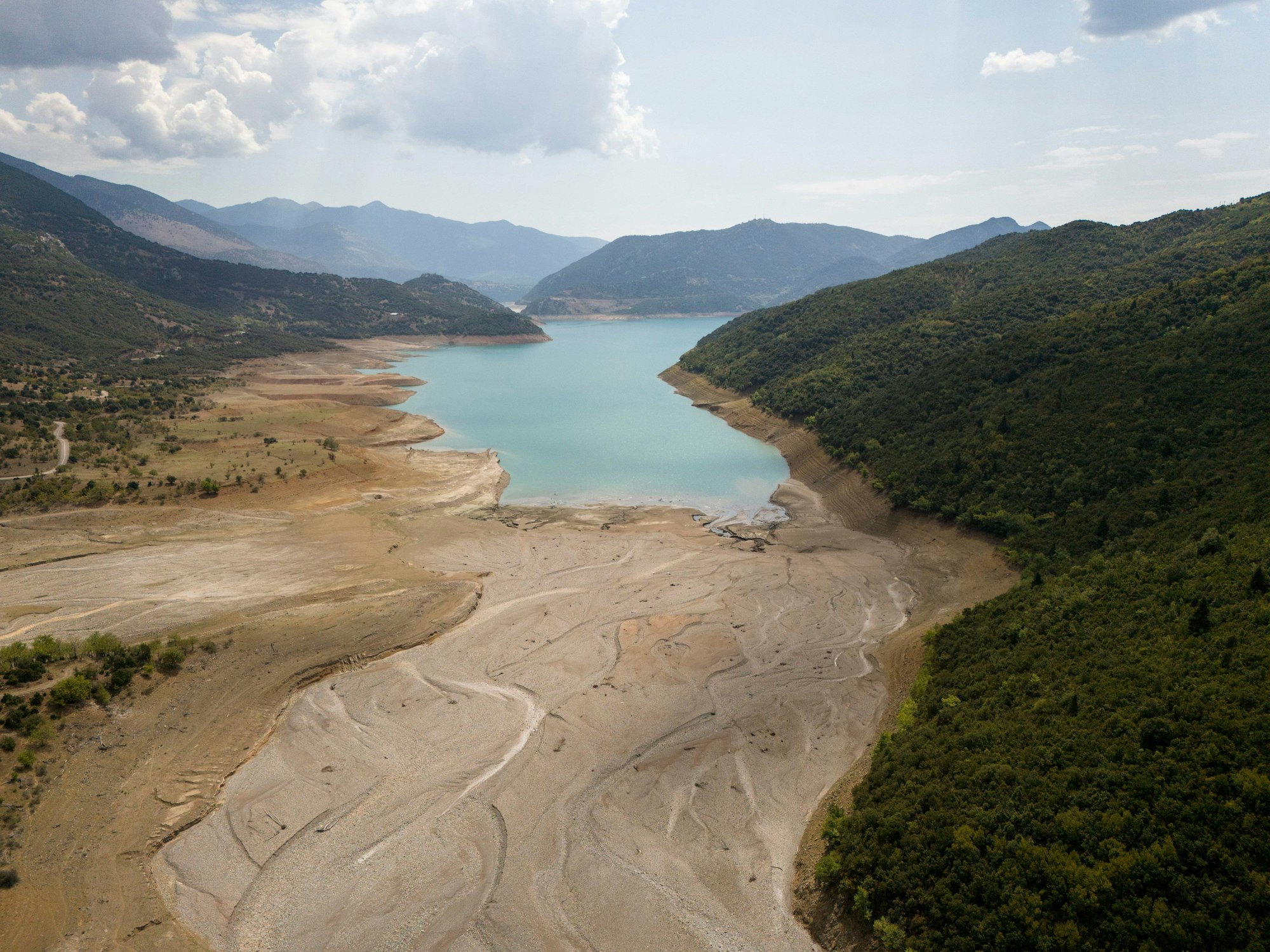Der niedrige Wasserstand des Mornos-Stausees ist deutlich zu sehen. Vor einem Bereich mit Wasser ist trockener Sand zu sehen, der sich von der Umgebung absetzt.