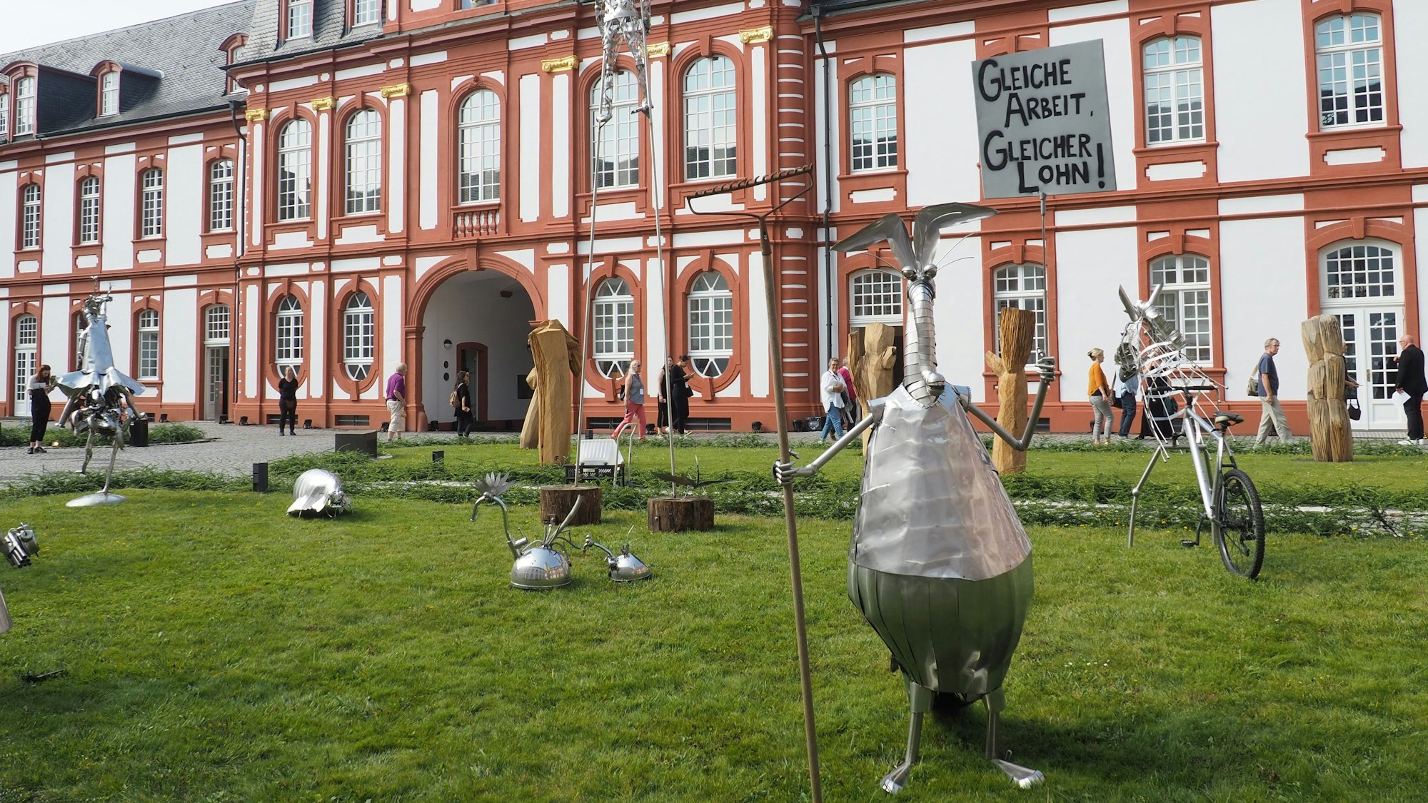 Vor der Abtei in Pulheim-Brauweiler stehen verschiedene kleine Skulpturen aus Blech. Vorne im Bild etwa ein stehender Ameisenbär, der eine Harke hält wie einen Speer.