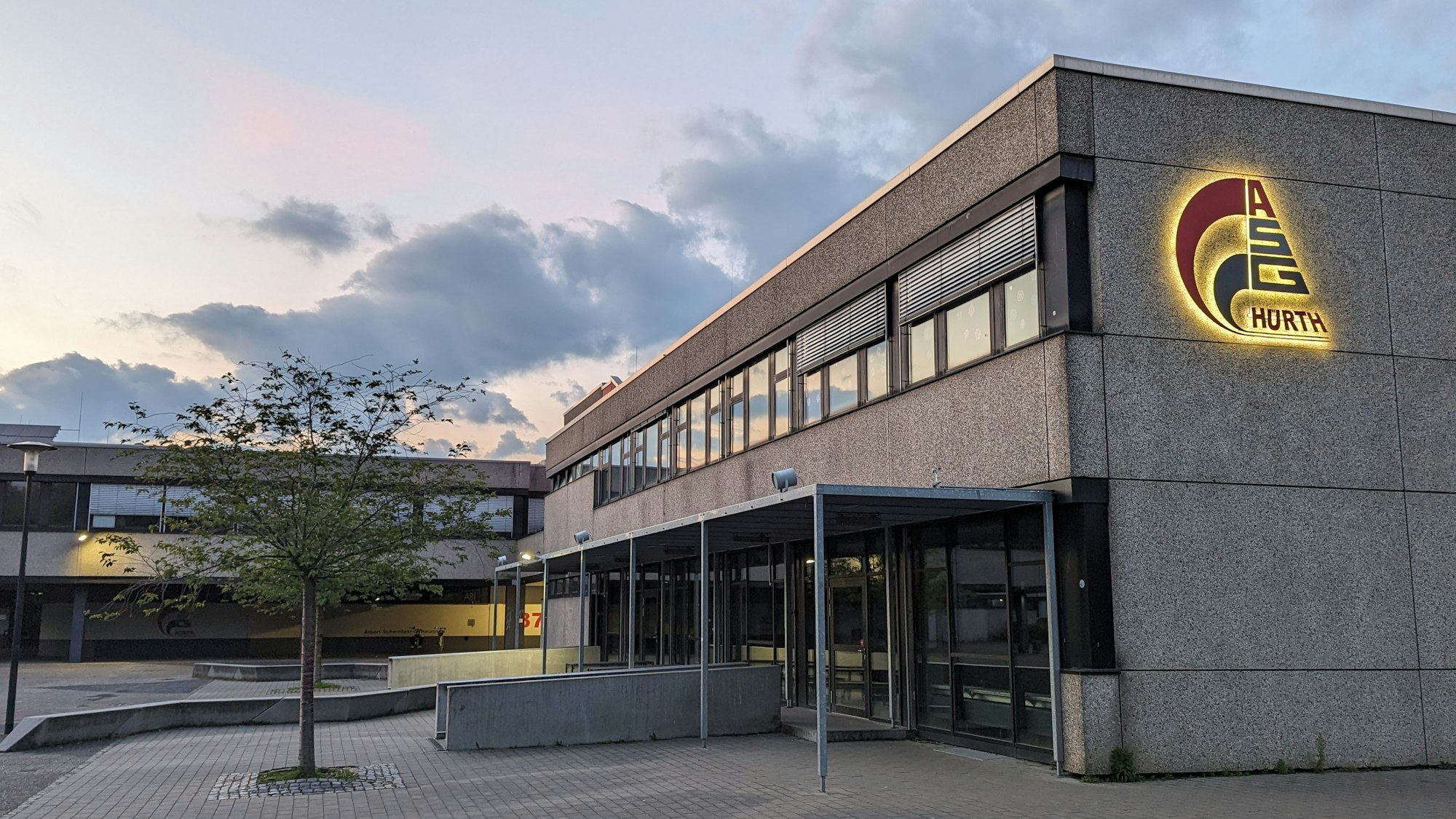 Das Bild zeigt eine Schule. An der Seite ist ein Logo mit der Aufschrift ASG Hürth. Vor der Schule steht ein kleiner Baum.