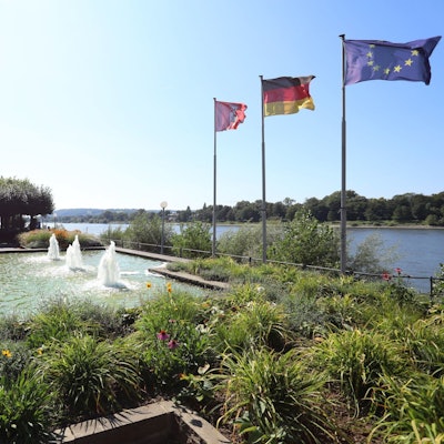Radfahrer fahren an einem sprudelnden Brunnen vorbei, rechts am Rheinufer wehen Flaggen im Wind.