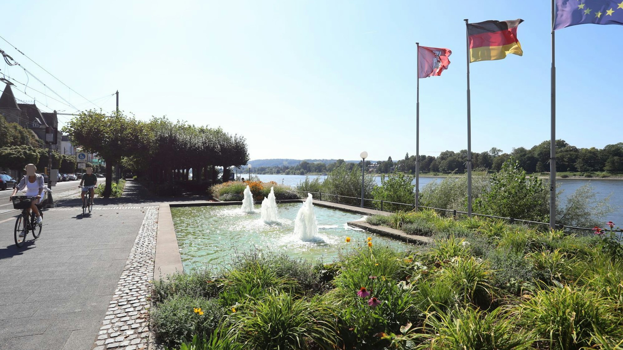 Radfahrer fahren an einem sprudelnden Brunnen vorbei, rechts am Rheinufer wehen Flaggen im Wind.