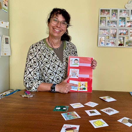 Eine Frau mit Brille zeigt grinsend auf ein Zeichentrick-Bild. Im Hintergrund hängen Fotos in einer Collage an einer Wand.
