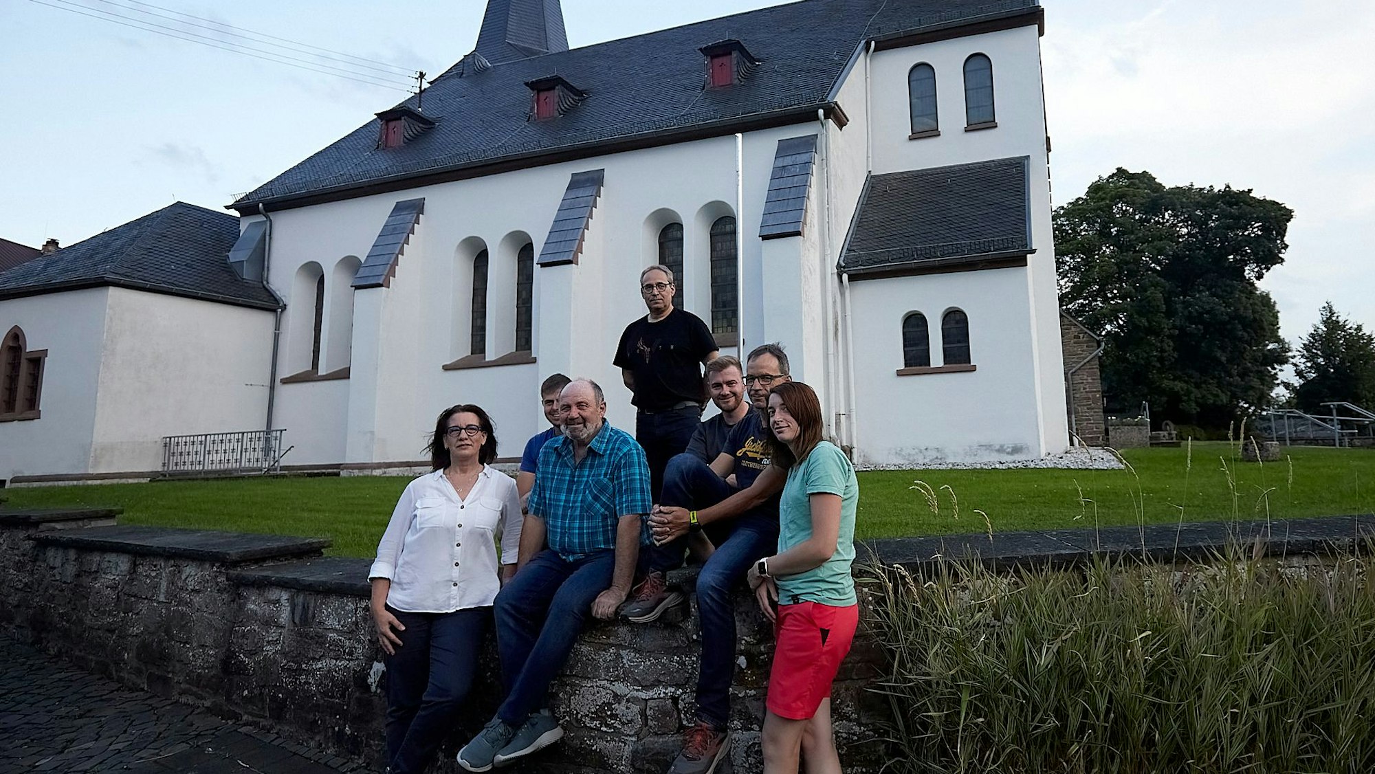 Eine Gruppe von sieben Frauen und Männern steht an einer Mauer, im Hintergrund eine Kirche.