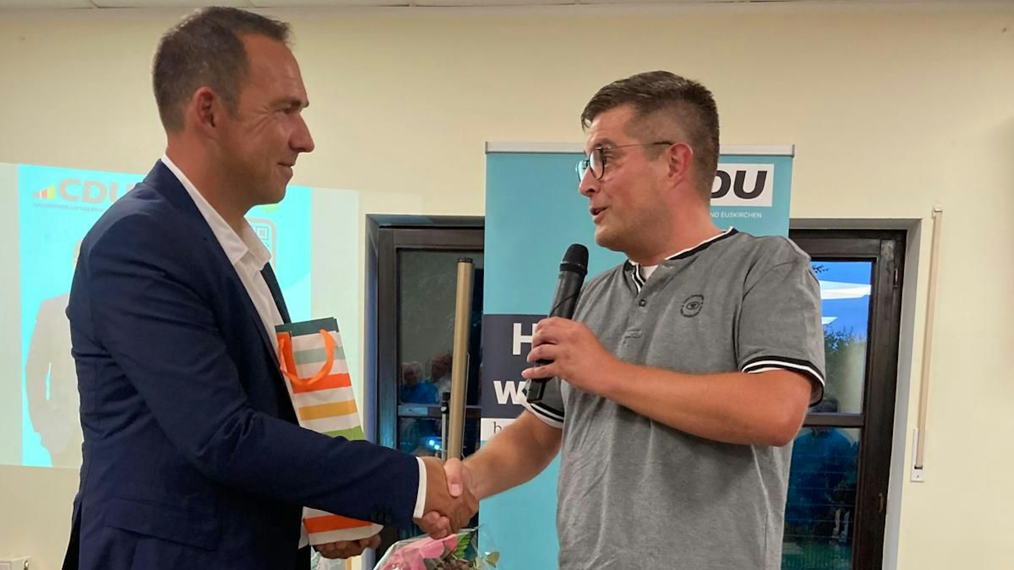 Ein Mann in Shirt und Jeans schüttelt einem Mann im Anzug die Hand, während er in ein Mikrofon spricht.