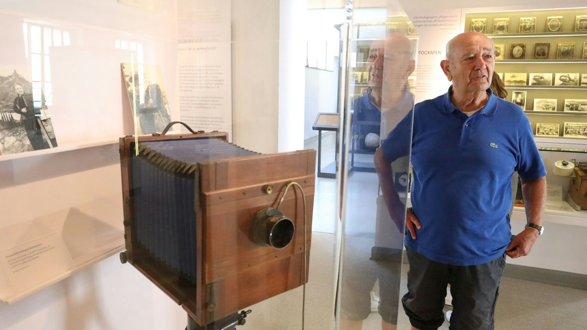 Ein Mann in blauem Poloshirt neben einer historischen Plattenkamera, die in einer  Vitrine steht.