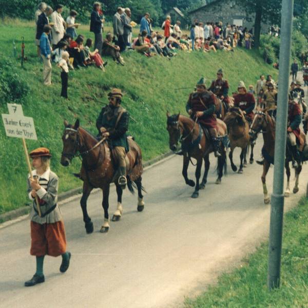 Männer in historischen Kostümen reiten auf einer schmalen Straße.