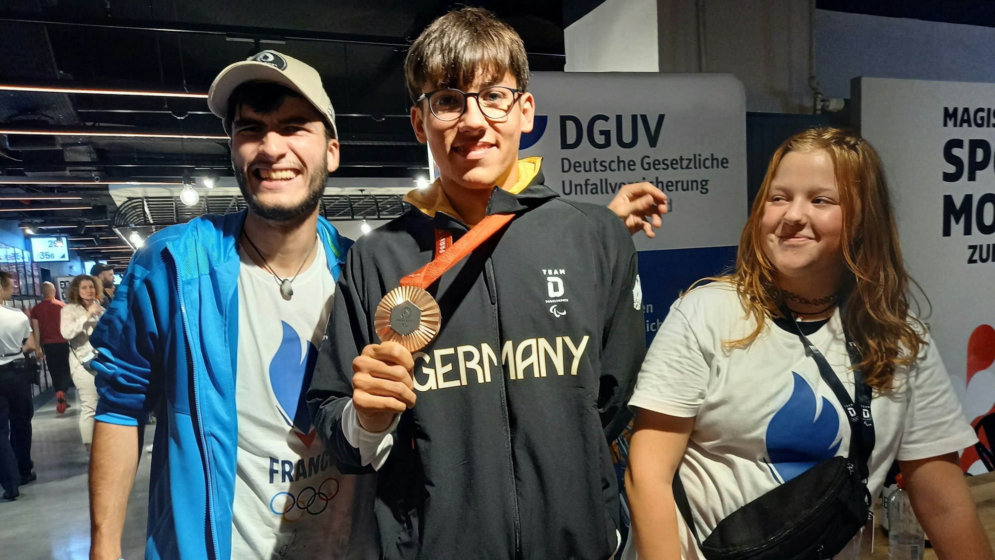 Zwei Schüler stehen mit dem Paraschwimmer und Bronzemedaillengewinner Maurice Wetekam zusammen. Der Sportler hält seine Bronzemedaille in der Hand.