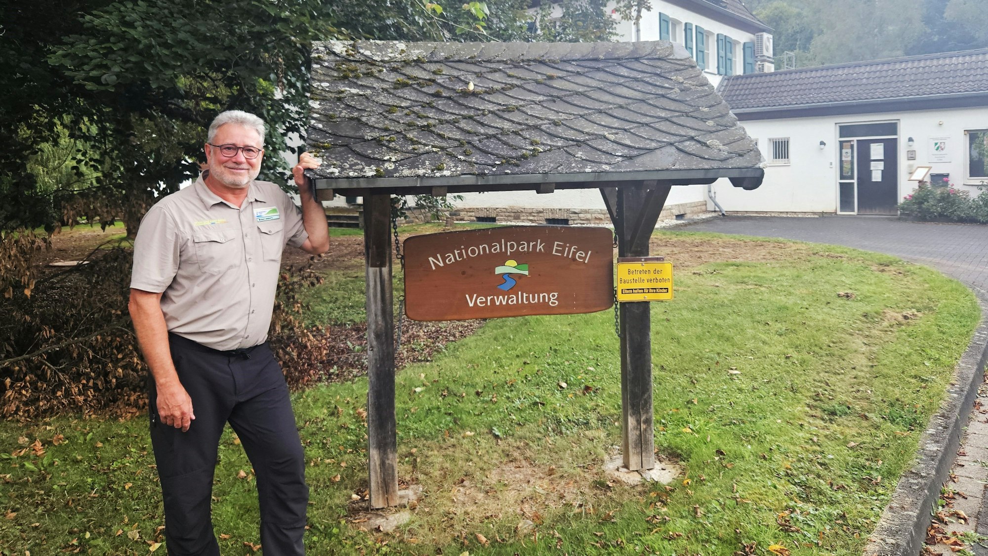 Ein Mann mit Brille steht neben einem Eingangsschild, auf dem „Nationalpark Eifel Verwaltung“ steht.