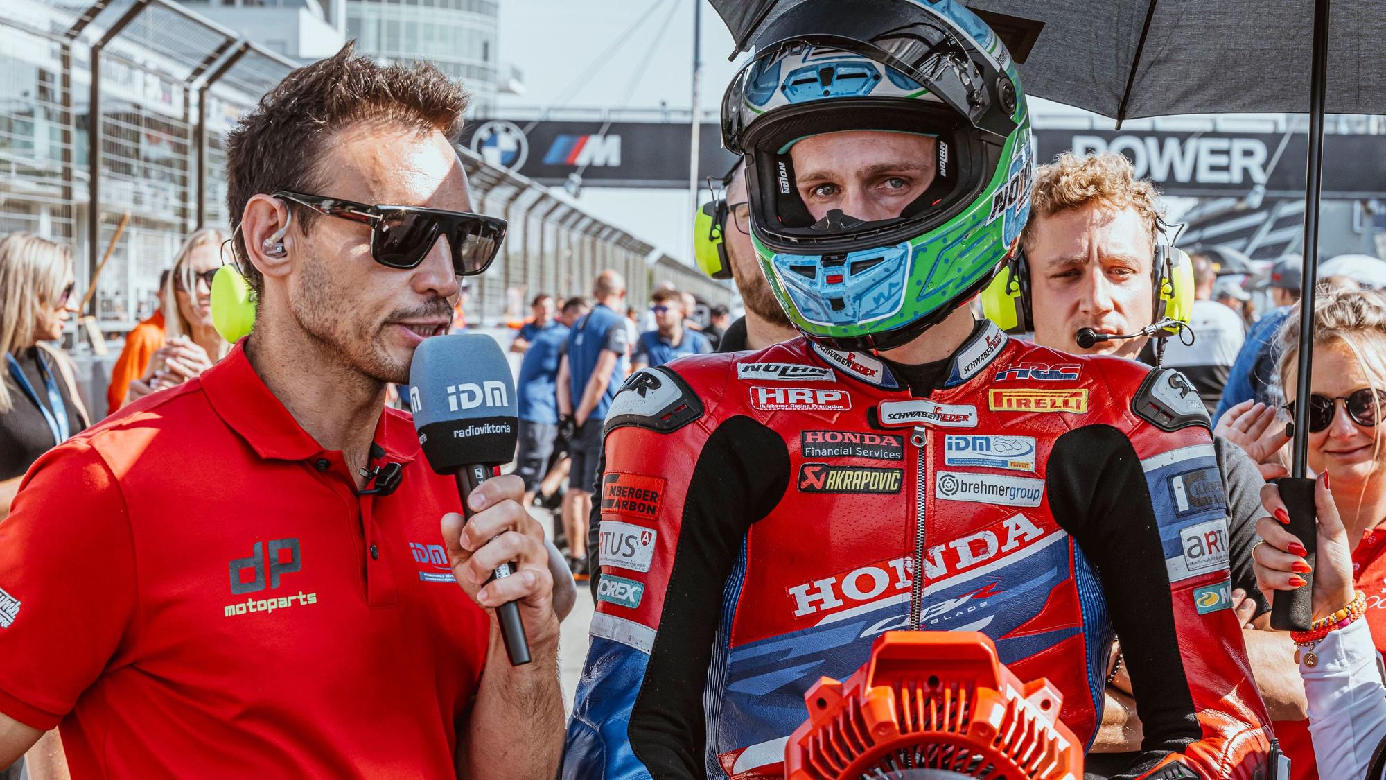 Zu sehen ist, wie ein Reporter einen Motorradrennfahrer per Mikrofon interviewt.
