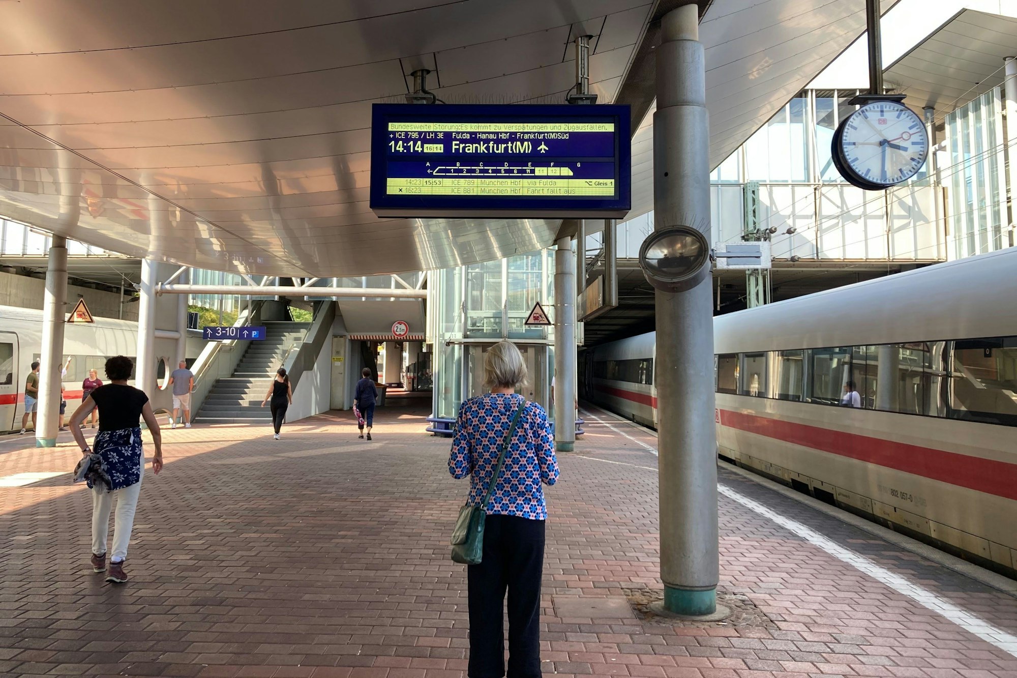 Eine Reisende steht auf dem Bahnsteig und schaut sich eine Anzeige der Deutsche Bahn mit dem Hinweis auf Verspätung an.