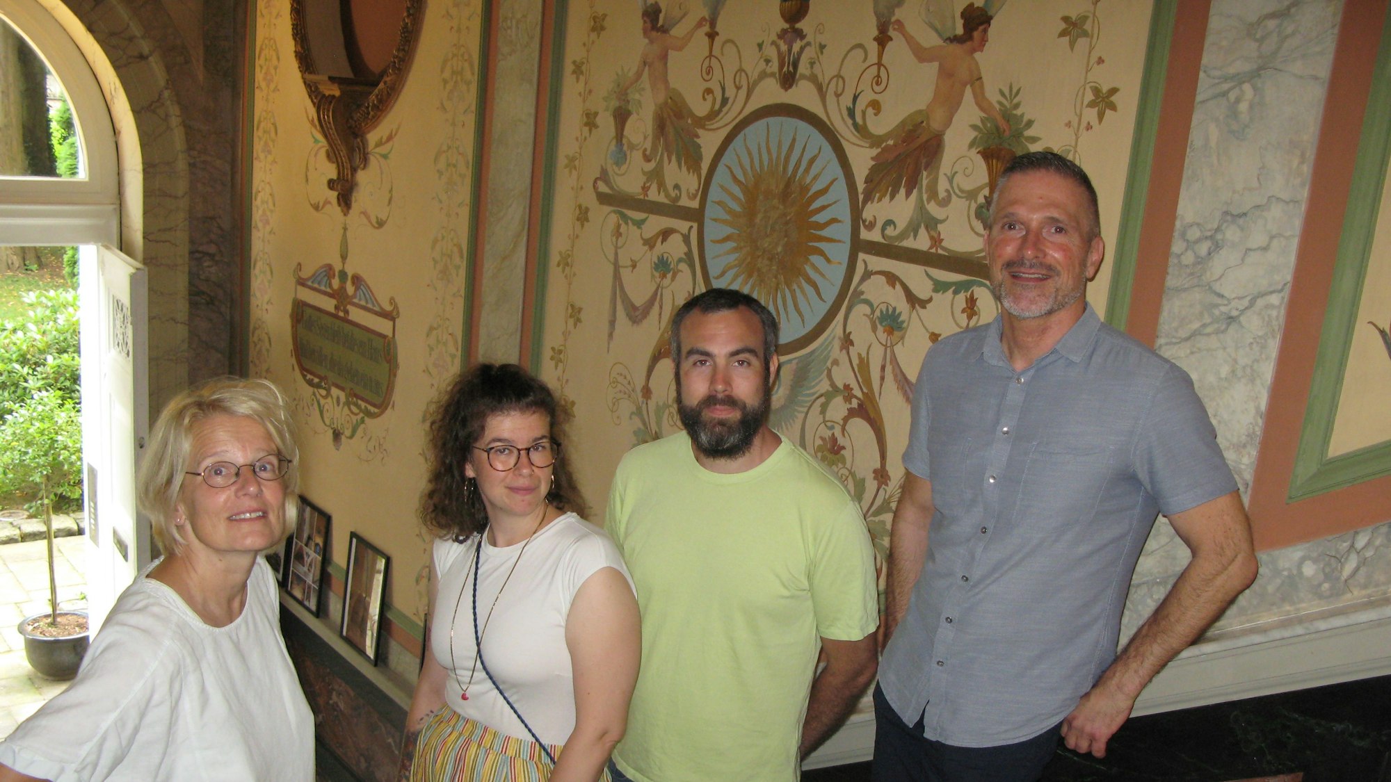 Zwei Frauen und zwei Männer stehen in einem Treppenhaus mit alten Wandmalereien.