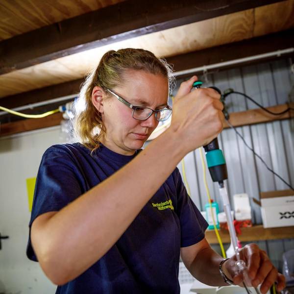 Eine Frau in einem dunkelblauen T-Shirt füllt mit einer Pipette Wasser in ein kleines Glas.