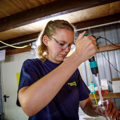 Eine Frau in einem dunkelblauen T-Shirt füllt mit einer Pipette Wasser in ein kleines Glas.