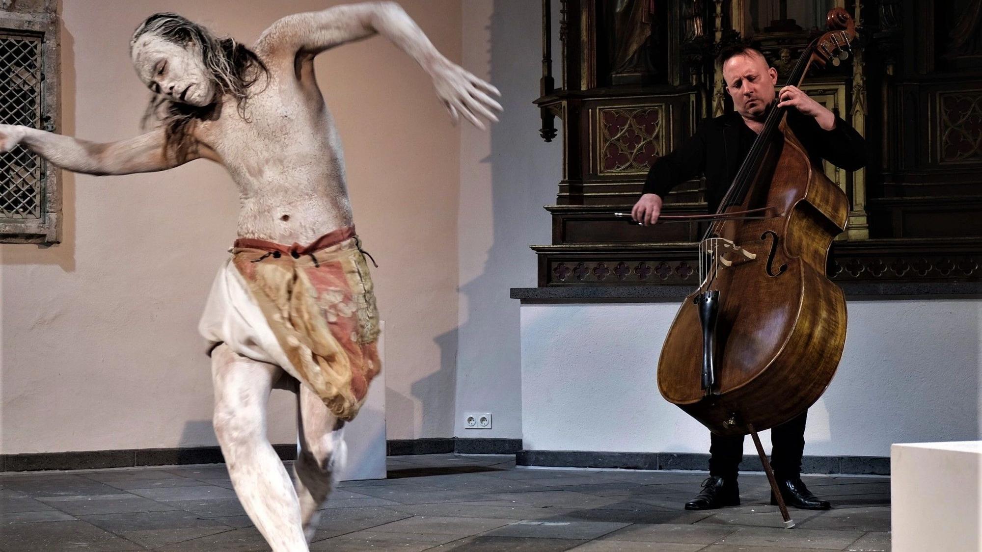 Ein Tänzer mit weiß geschminktem Körper und ein Kontrabassist in einer Kirche.