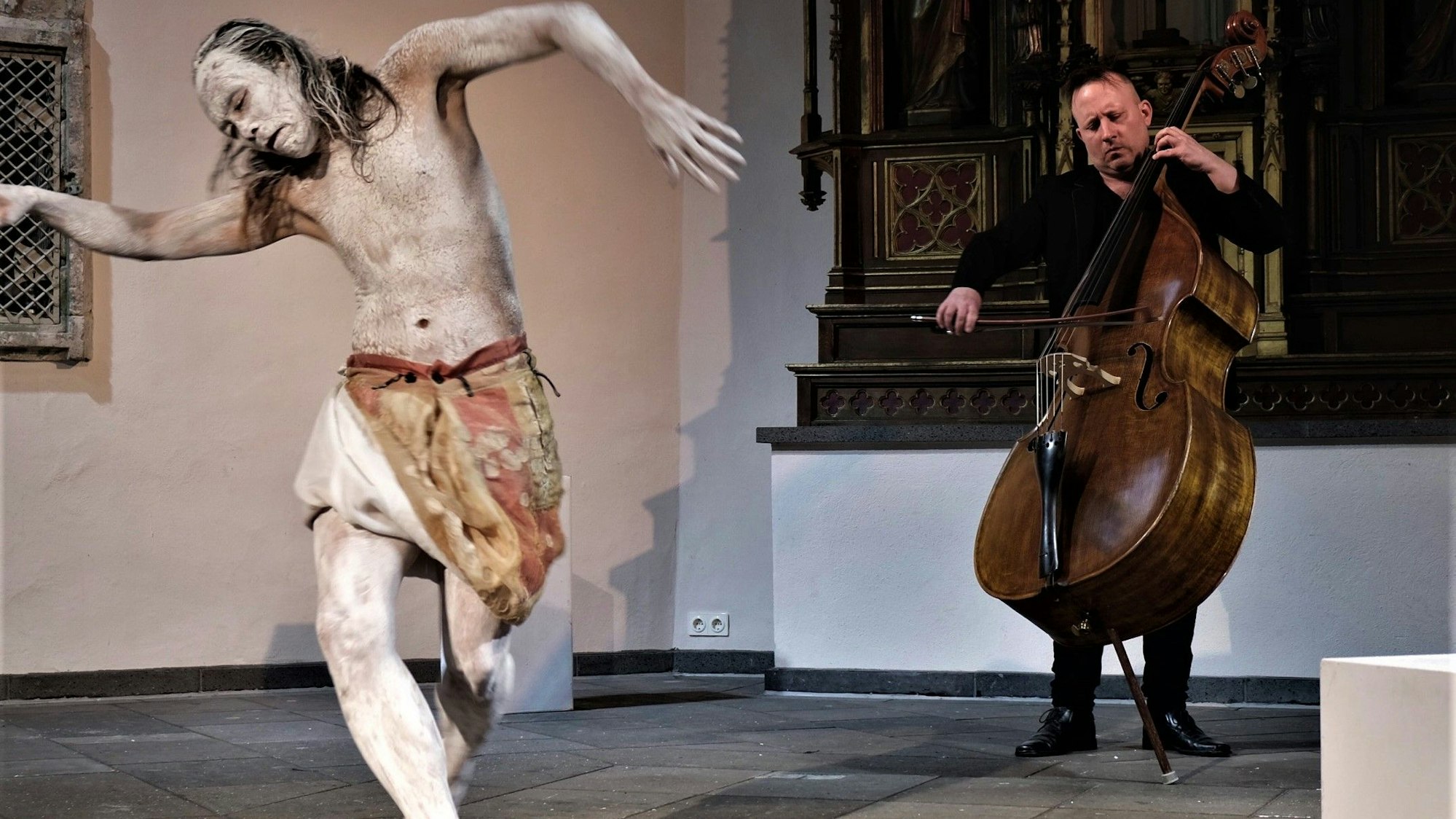 Ein Tänzer mit weiß geschminktem Körper und ein Kontrabassist in einer Kirche.