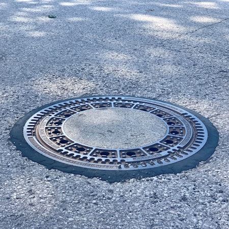 Zu sehen ist ein runder Gulli in einer Straße.