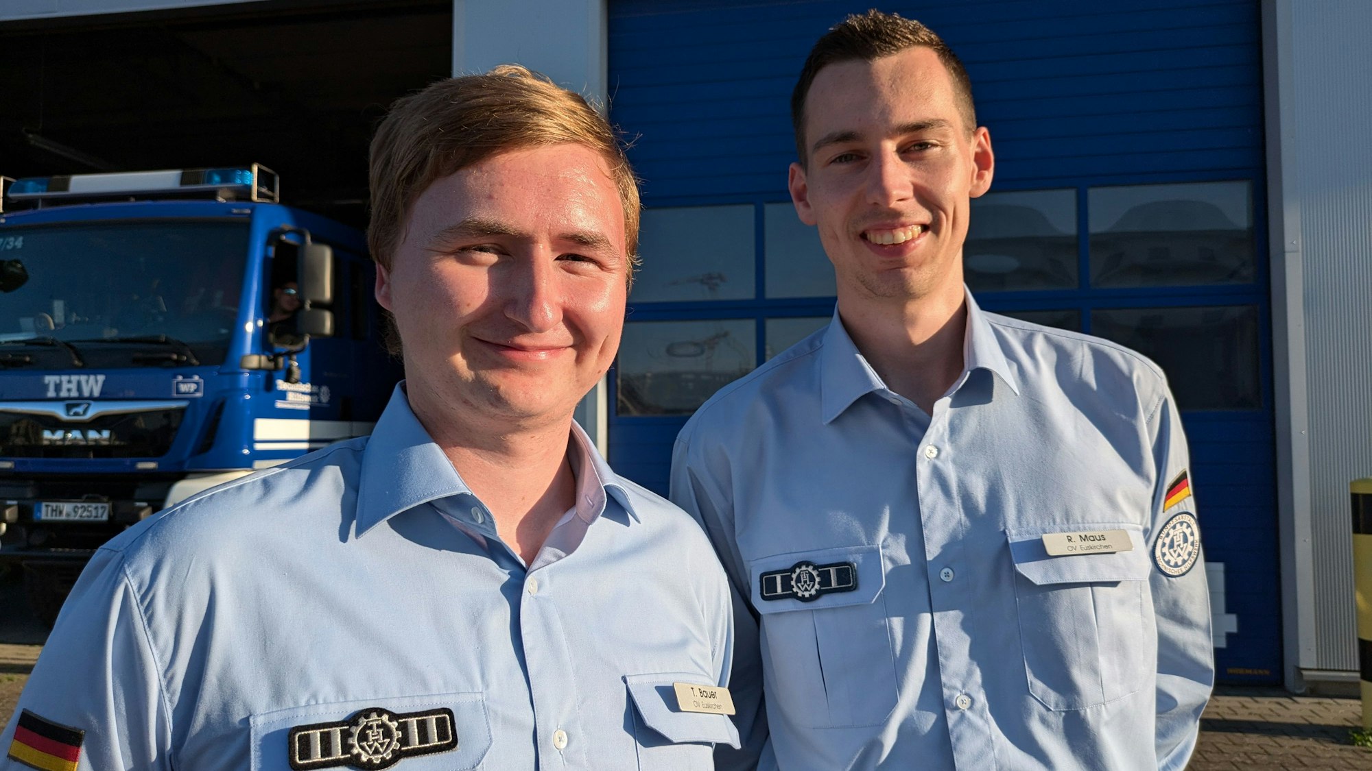 Tim Bauer und Raphael Maus stehen in hellblauem Uniform-Hemd vor einer THW-Garage.