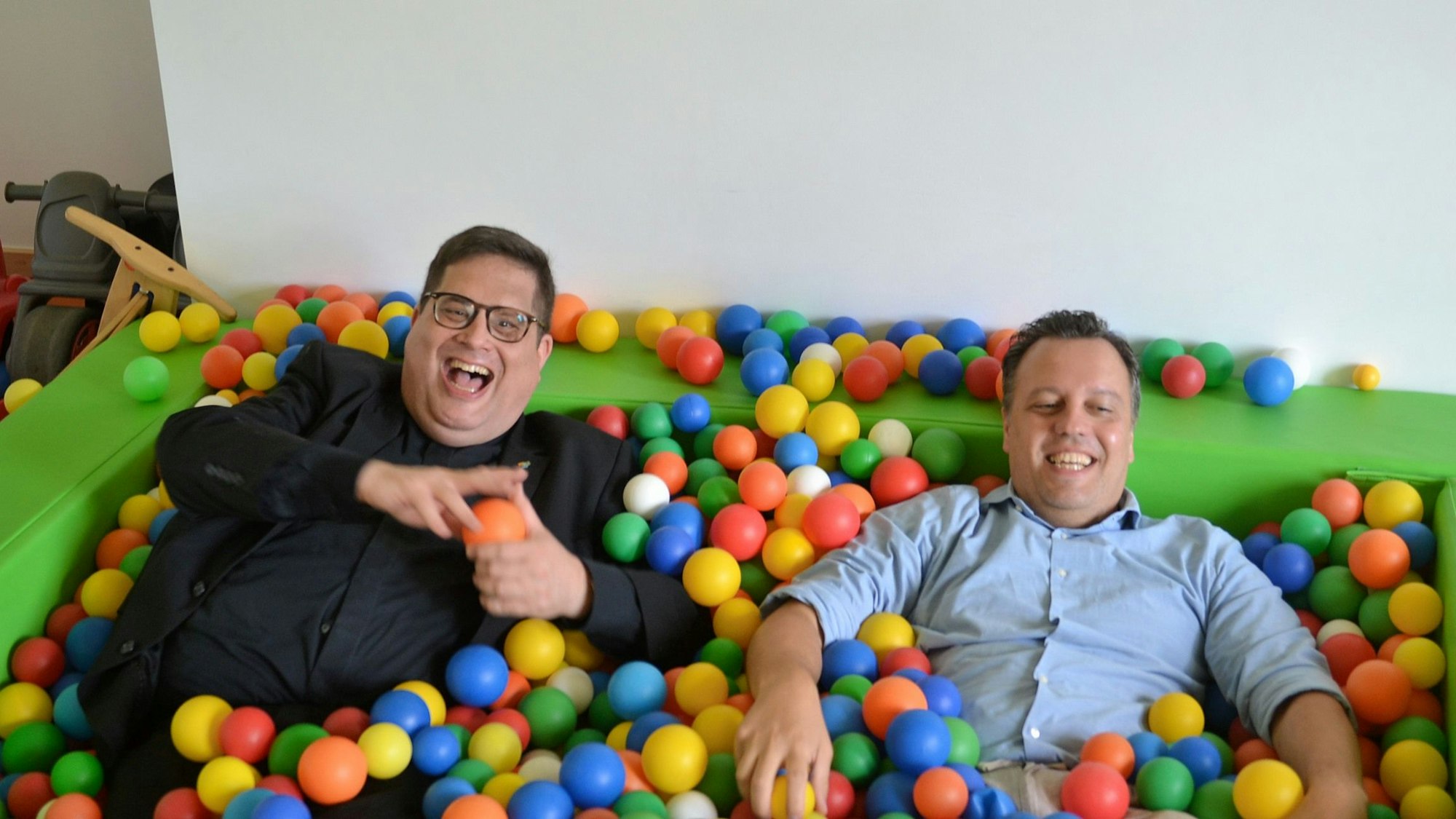 Zwei Männer liegen in einem Bällebad. Der rechte trägt schwarzes Hemd und schwarzes Sakko, der linke ein hellblaues Hemd.