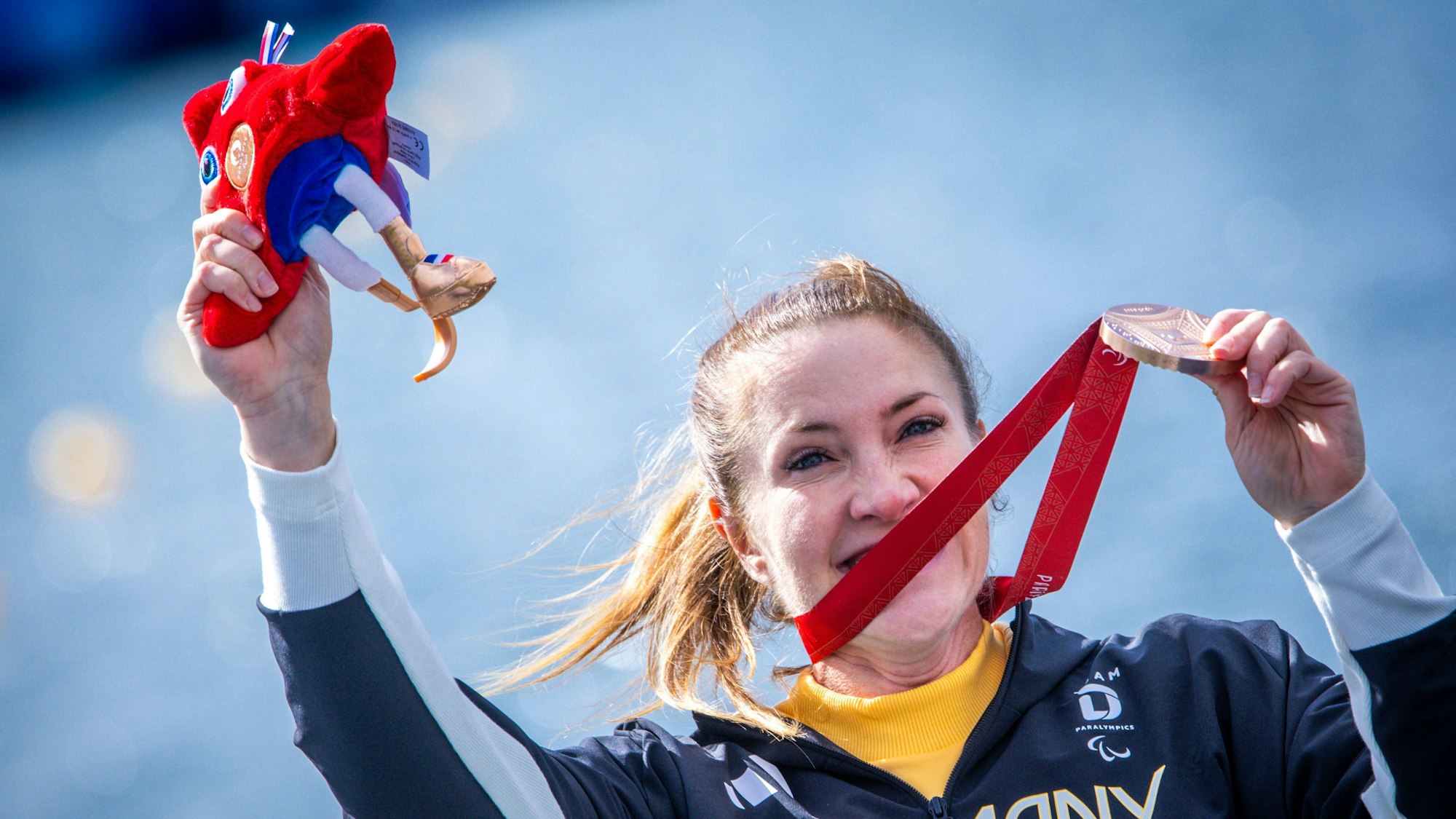 Edina Müller jubelt nach dem Rennen mit ihrer Bronzemedaille und hält Phryges, das Maskottchen der Paralympics, in ihrer Hand.