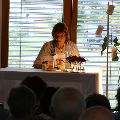 Eine Frau mit Brille sitzt an einem Tisch mit einer Leselampe und liest aus einem Buch vor.