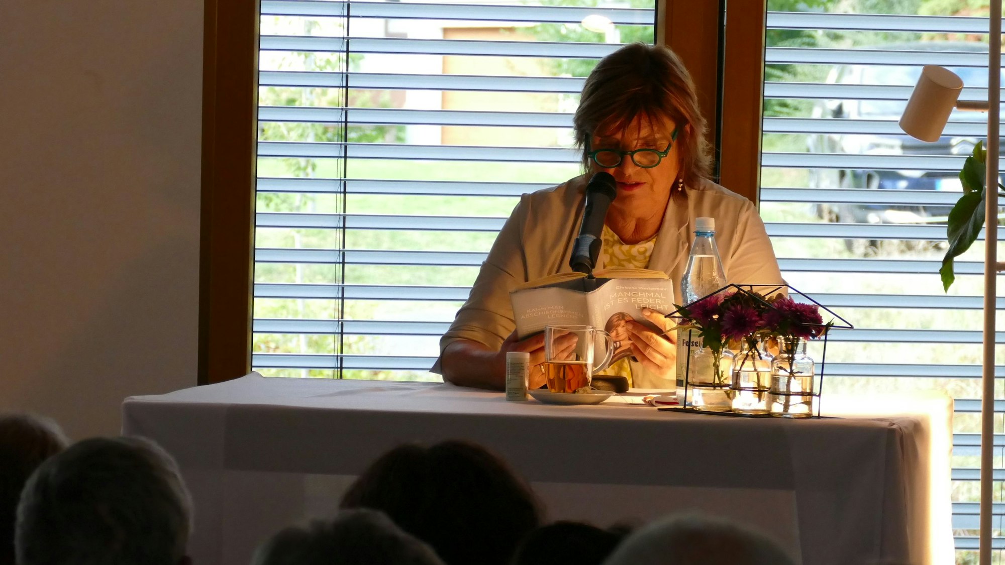 Eine Frau mit Brille sitzt an einem Tisch mit einer Leselampe und liest aus einem Buch vor.