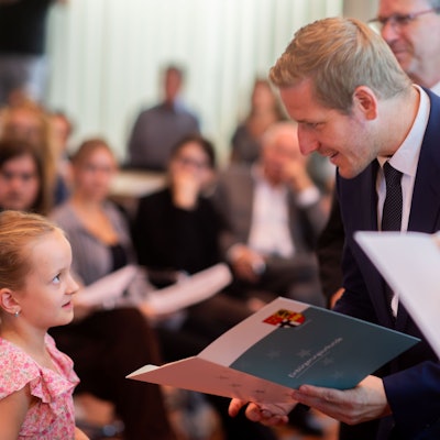 Markus Ramers (rechts im Bild) überreicht in einem dunkelblauen Anzug einem Mädchen mit rosa-geblümtem Oberteil und blonden Haaren eine Urkunde.