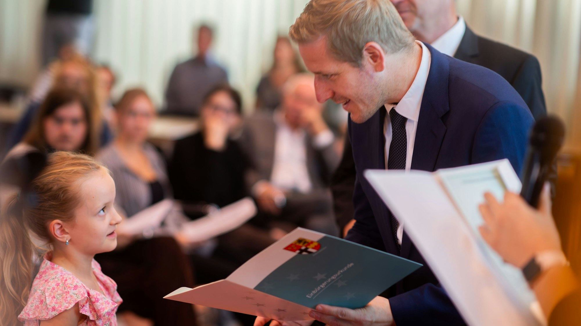 Markus Ramers (rechts im Bild) überreicht in einem dunkelblauen Anzug einem Mädchen mit rosa-geblümtem Oberteil und blonden Haaren eine Urkunde.