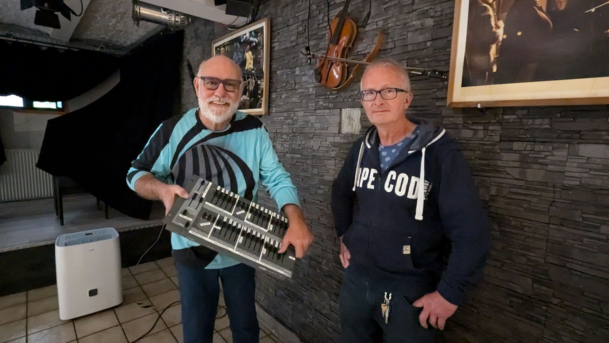 Zwei Männer zeigen ein Mischpult im Jazzkeller. Im Hintergrund ist ein Trocknungsgerät zu sehen.