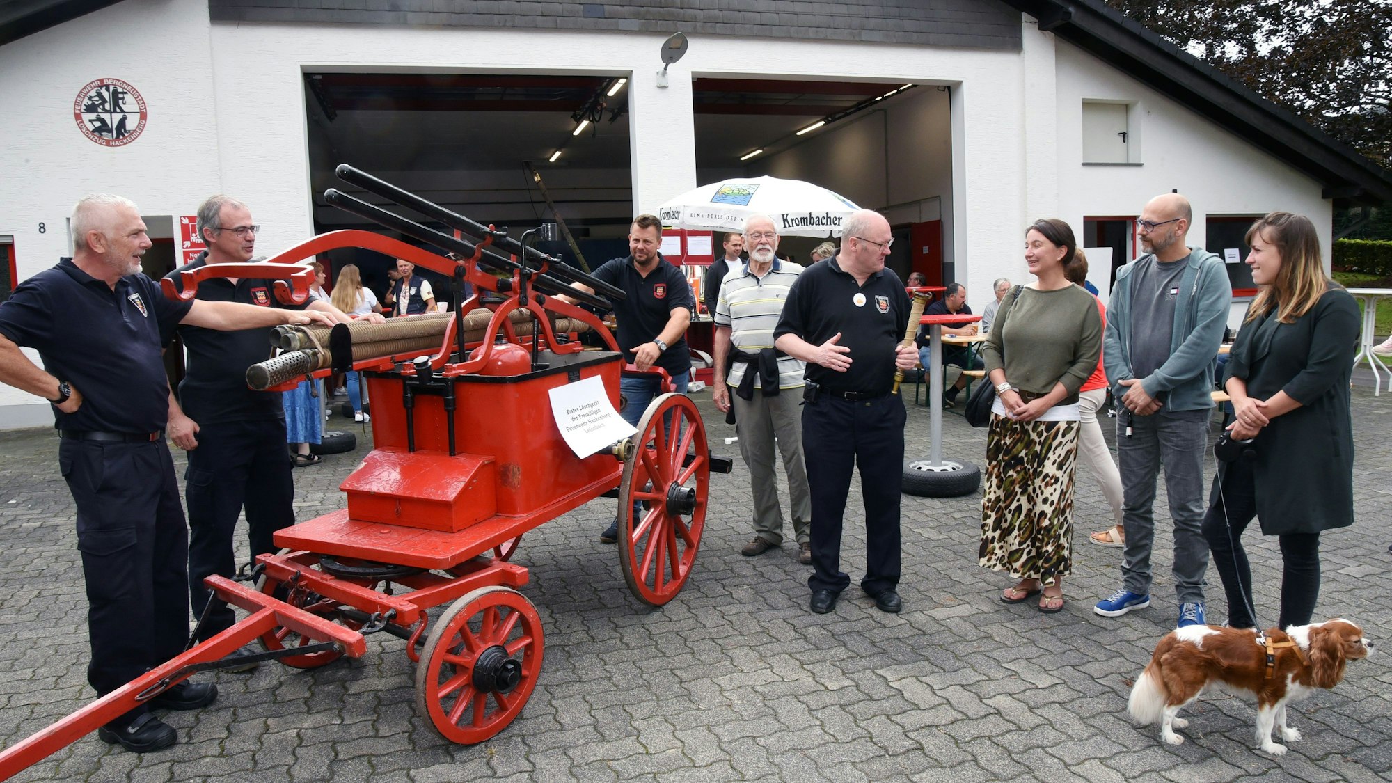 Feuerwehrleute zeigen ihren Besuchern eine fahrbare Wasserspritze auf Holzrädern.