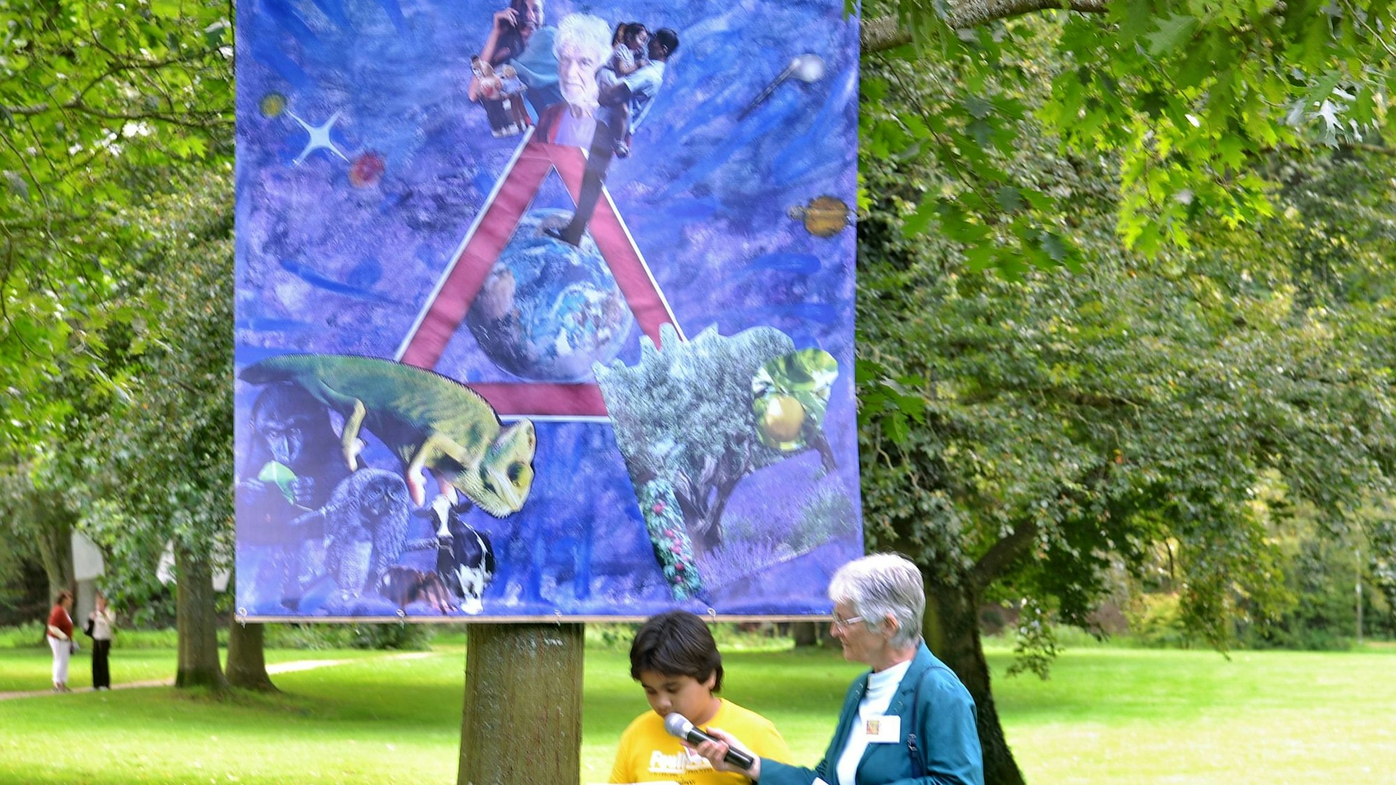 Der junge Künstler erläutert sein Bild, das an einem Baum hängt. Eine Frau hält das Mikrofon, in das er spricht.
