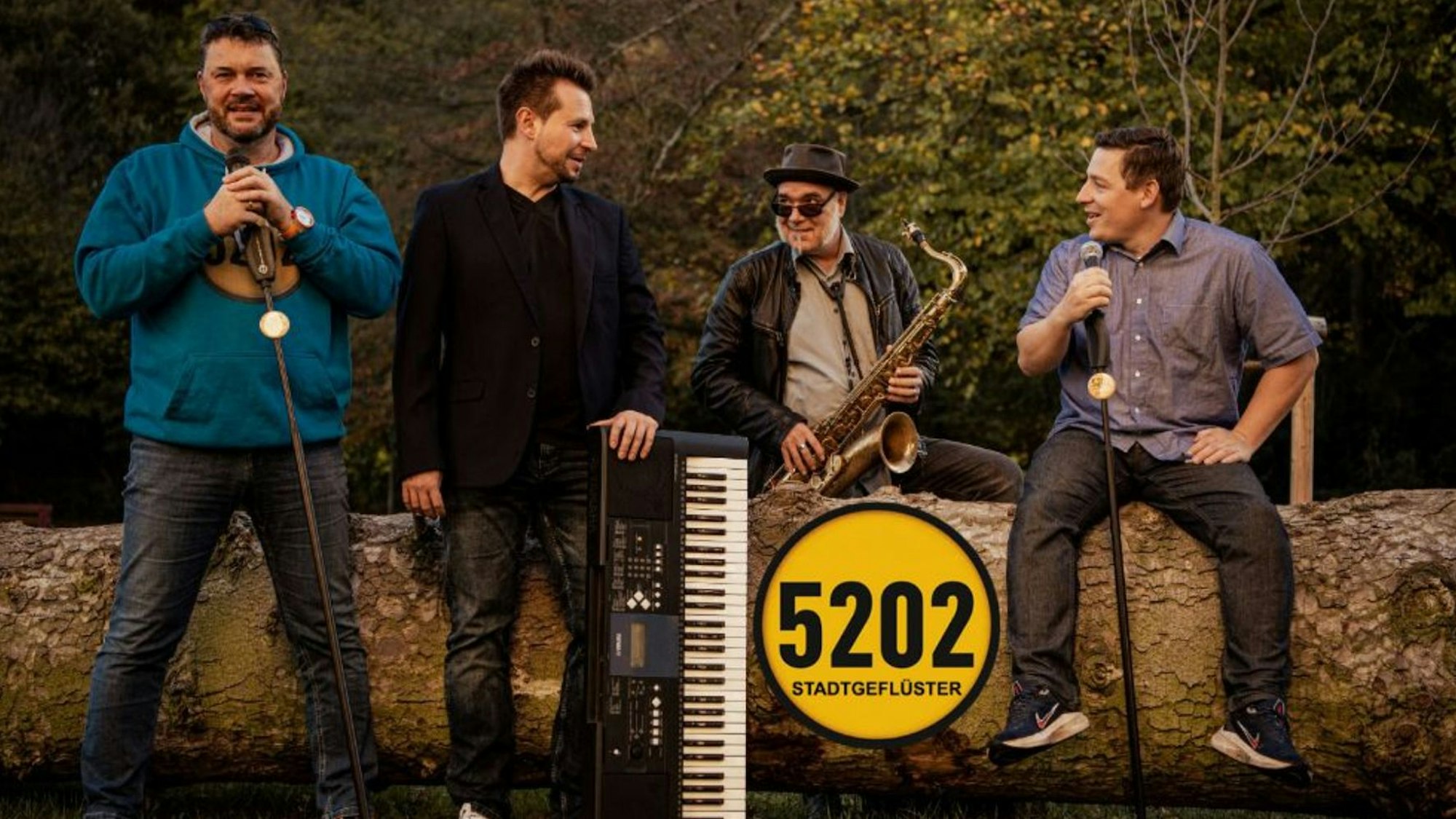 Die vier Mitglieder der Hennefer Band 5202 mit Musikinstrumenten an einem quer liegenden Baumstamm.