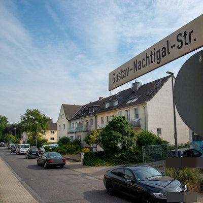 Ein Straßenschild und die dazugehörige Straße mit Reihenhäusern und parkenden Autos ist zu sehen.