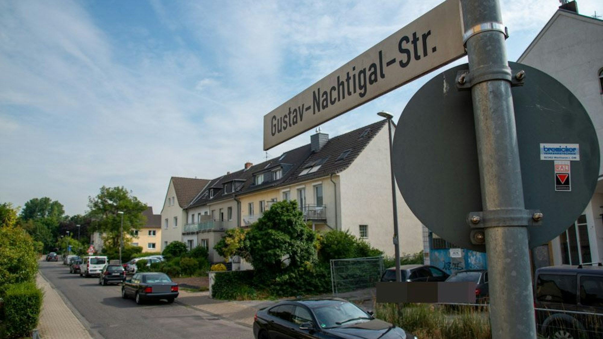 Ein Straßenschild und die dazugehörige Straße mit Reihenhäusern und parkenden Autos ist zu sehen.
