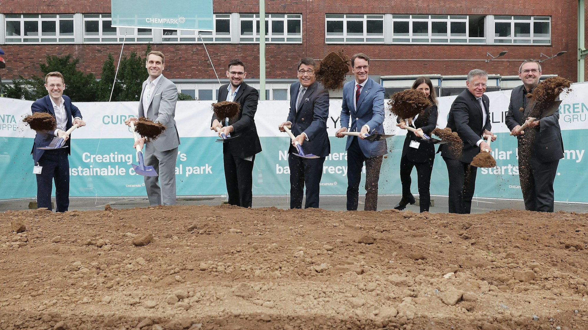 Acht Personen halten einen Spaten in der Hand und werfen damit Erde in die Höhe.