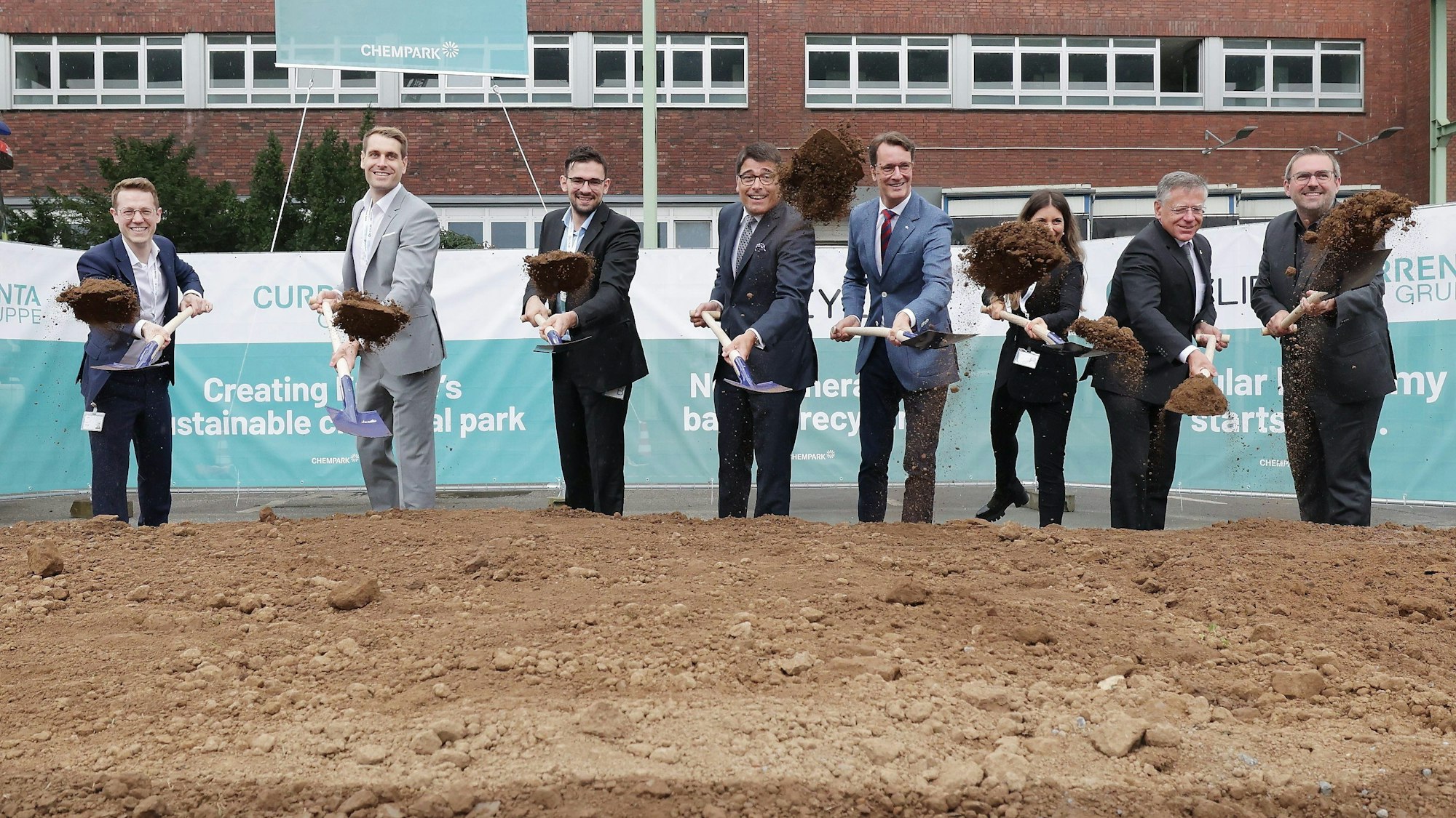 Acht Personen halten einen Spaten in der Hand und werfen damit Erde in die Höhe.