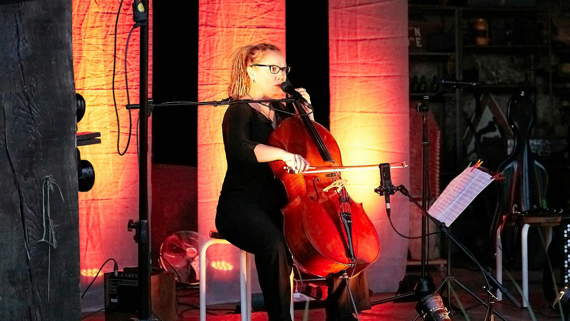 Die Musikerin sitzt, mit Licht stimmungsvoll in Szene gesetzt, auf einem Stuhl, vor sich ihr Cello und ein Mikrofon.
