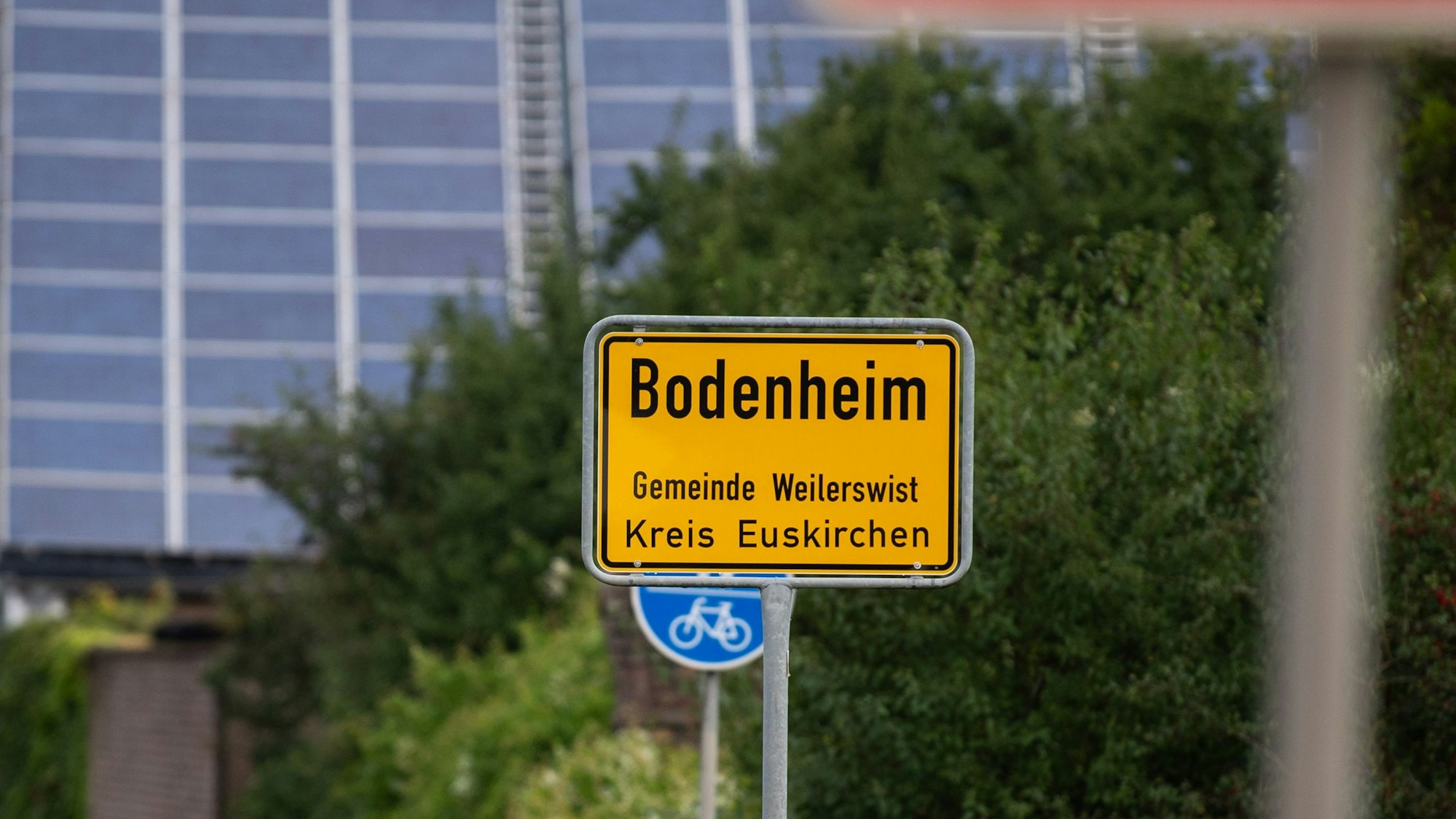 Ein gelbes Ortsschild mit der Aufschrift: „Bodenheim, Gemeinde Weilerswist, Kreis Euskirchen“ steht vor grünen Büschen.