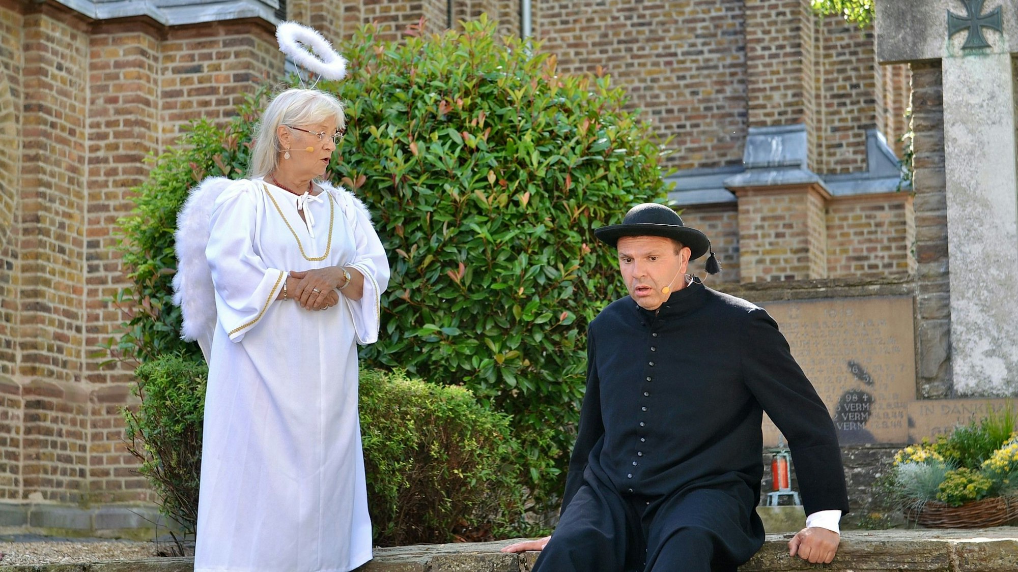 Helga Hettmer ist als Engel verkleidet, Thomas Kreuz als Priester. Im Hintergrund die Rövenicher Kirche.